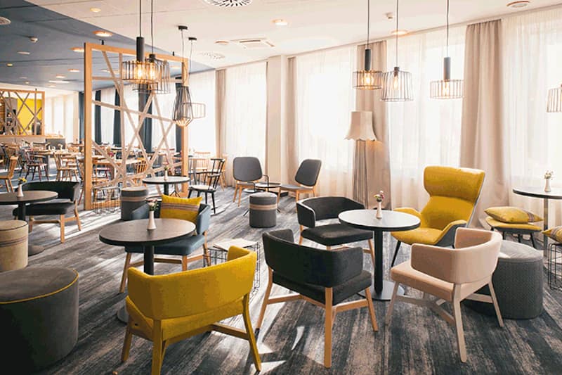 Helle Hotellounge mit verschiedenen Stuhlgruppen, kleinen Tischen und Hängelampen vor großen Fenstern.