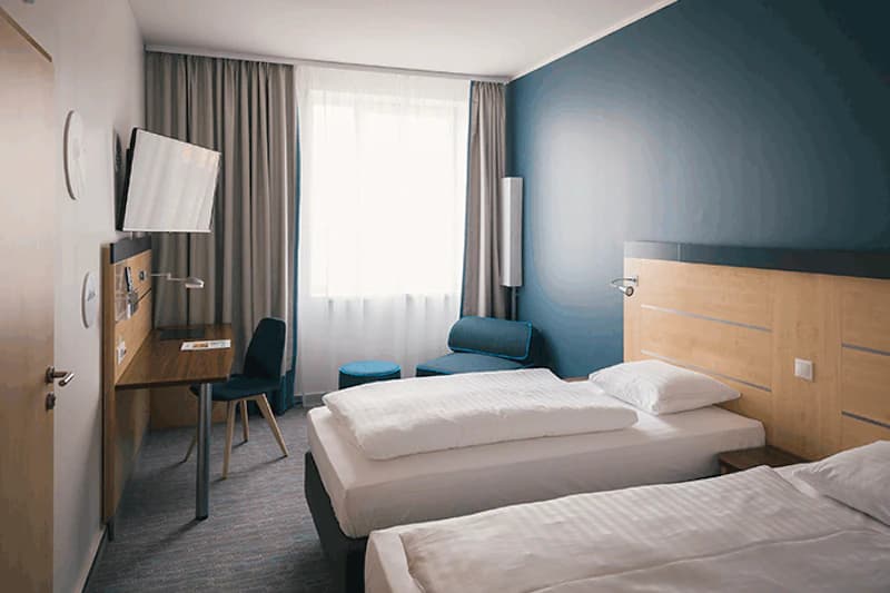 Hotelzimmer mit zwei Einzelbetten, Holzkopfteil, Schreibtisch, Fernseher, Sitzgelegenheit und Fenster mit Vorhang.