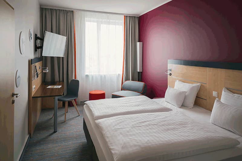 Zimmer mit zwei Betten, burgunderrote Akzentwand, Schreibtisch mit Wandfernseher, Sessel und Vorhangfenster.