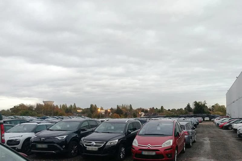 Großer Außenparkplatz mit Reihen geparkter Autos unter bedecktem Himmel und Gebäuderand rechts.