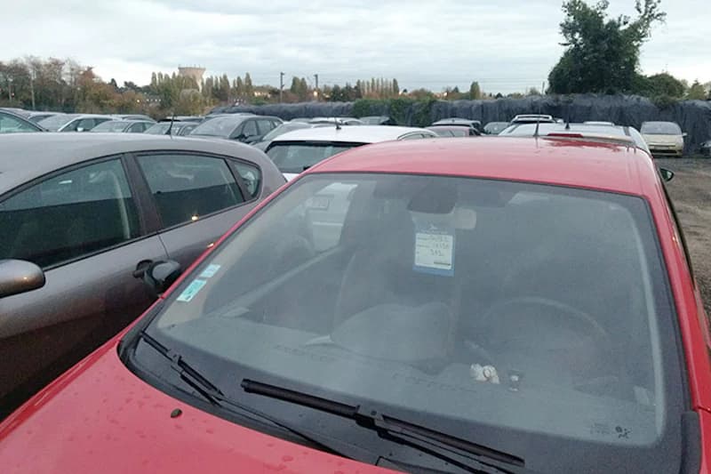 Gezaeunter Parkplatz mit Reihen geparkter Autos, rotes Fahrzeug im Vordergrund mit Zettel am Innenspiegel.
