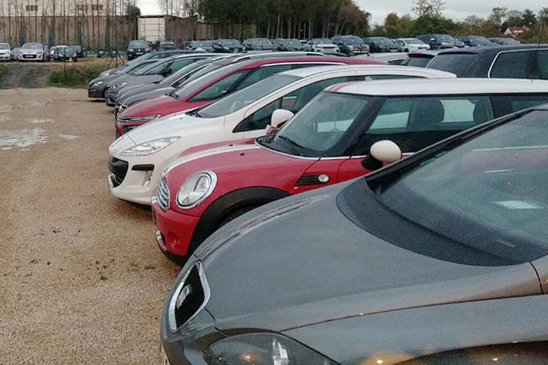 Reihe geparkter Autos auf einem kiesigen Parkplatz vor einem Zaun und Baumreihe.
