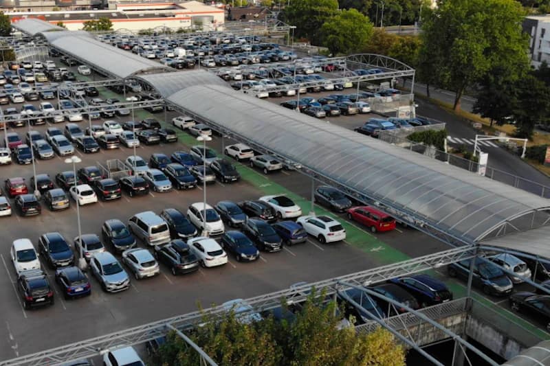 Großer Außenparkplatz mit zahlreichen Autos und überdachten Fußwegen, Beleuchtung und markierten Stellreihen.