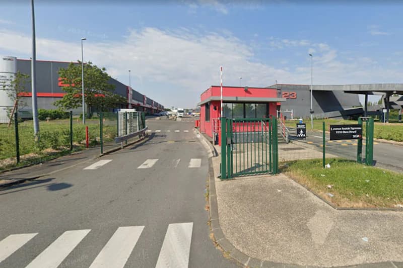 Grünes Gittertor und rotes Pförtnerhäuschen an Zufahrt mit Zebrastreifen vor Lagerhalle mit Zahl 23.