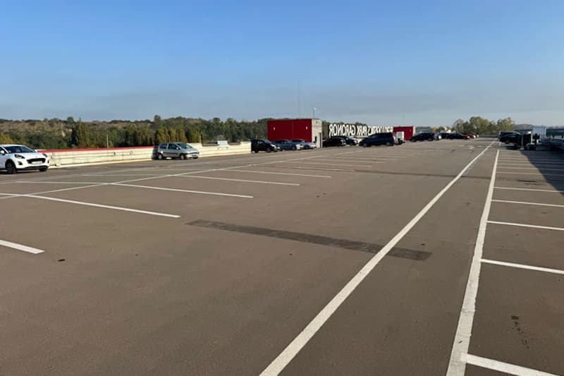 Großer oberirdischer Parkplatz mit Parkmarkierungen, wenigen Autos, rotem Container mit weißer Schrift und Baumbestand im Hintergrund.