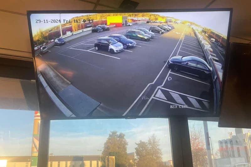 Monitor zeigt Parkplatz mit mehreren geparkten Autos und markierten Stellflächen sowie Zeitstempel oben links.