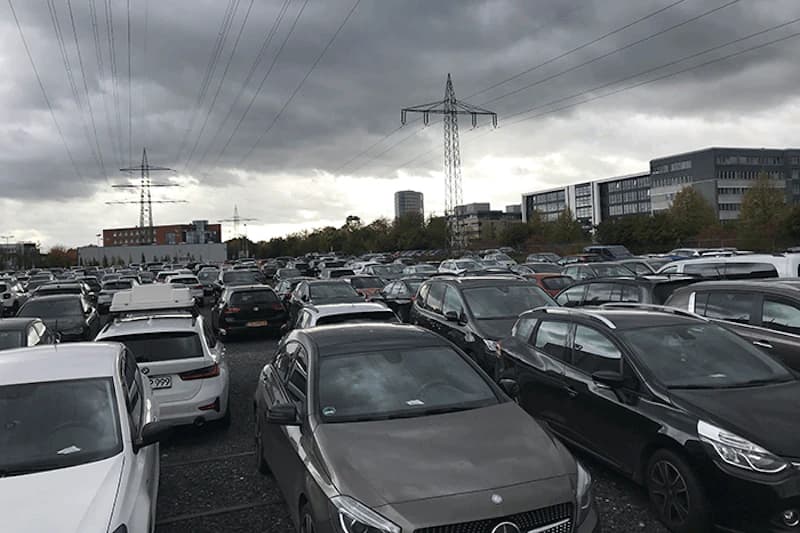 Großes, freies Parkplatzgelände mit dicht beparkten Reihen, Hochspannungsmasten und bewölktem Himmel.