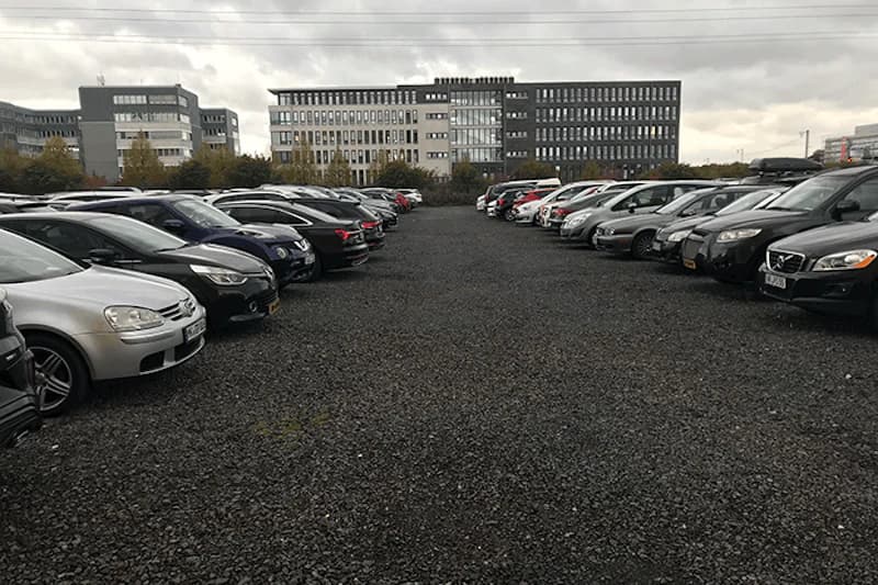 Schotterparkplatz mit zwei Reihen geparkter Autos und mehrstöckigen Gebäuden im Hintergrund bei bedecktem Himmel.