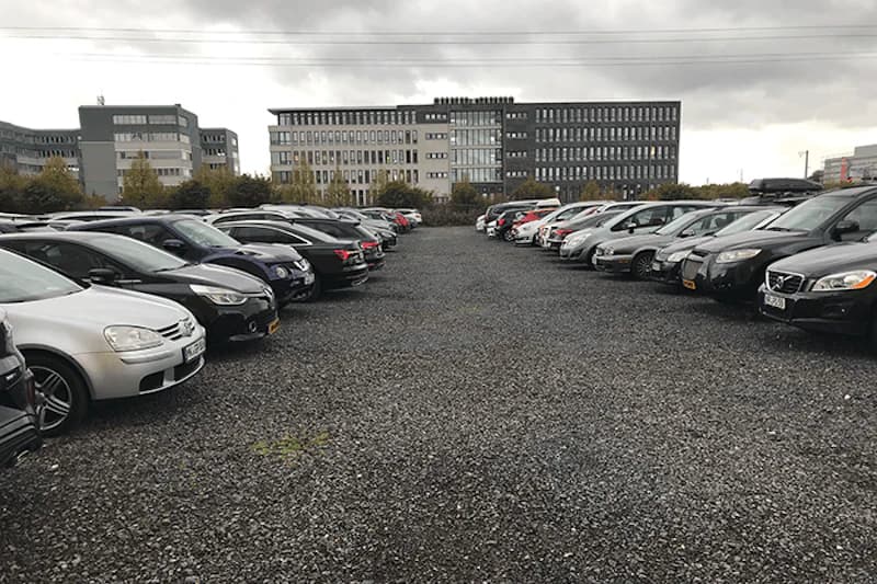 Schotterparkplatz mit zwei Reihen geparkter Autos und mehrstöckigem Bürogebäude im Hintergrund unter bewölktem Himmel.