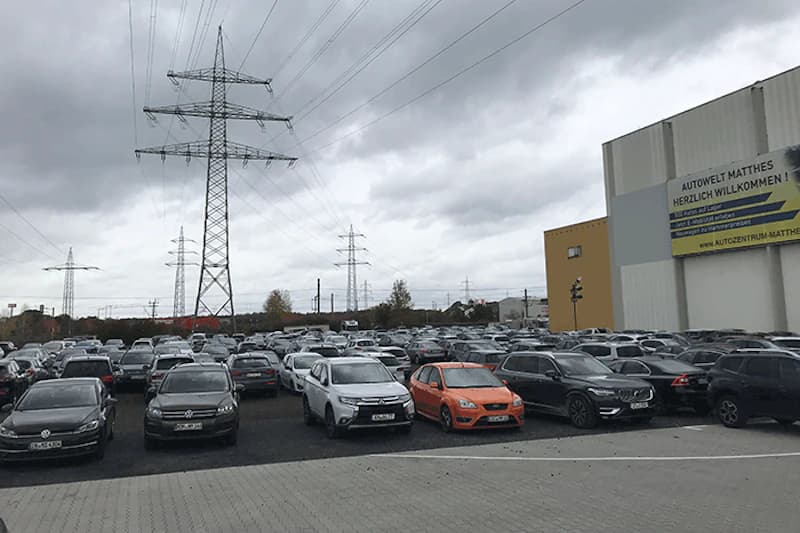 Großes Parkplatzfeld voller geparkter Autos unter Strommasten und bewölktem Himmel, Gebäude mit Werbeplakat rechts.