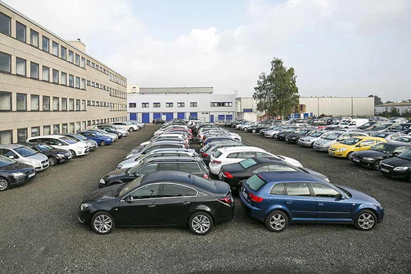 Mehrere Reihen geparkter Autos auf offenem Schotterparkplatz zwischen Bürogebäude und Hallen, einzelner Baum im Hintergrund.