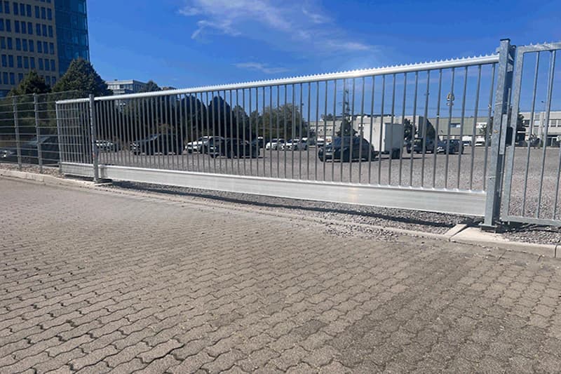Metallschiebetor und umzäunter Parkplatz mit geparkten Autos, gepflasterter Vorplatz und Gebäude im Hintergrund.
