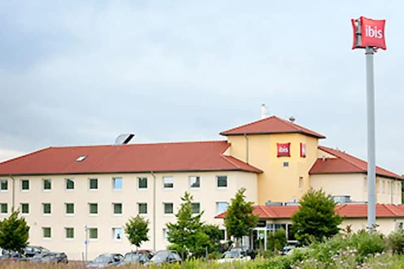 Dreistöckiges ibis Hotel mit rotem Dach, Parkplatz mit Pkw, kleinen Bäumen und hohem Schild mit ibis-Logo.