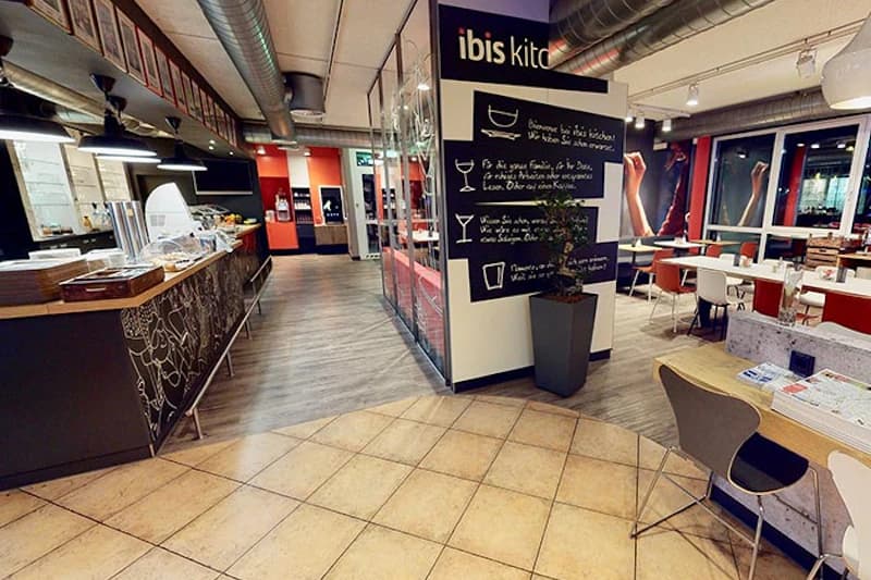 Hotelgastraum mit Theke, Sitzgruppen, Wandtafel und sichtbarem ibis kitchen Schriftzug.
