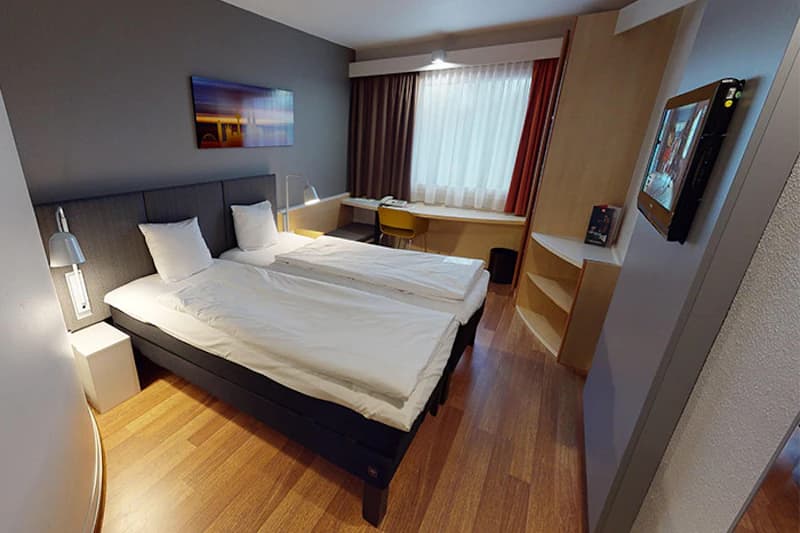 Hotelzimmer mit zwei Einzelbetten, Nachttischen und Lampen, Schreibtisch am Fenster und wandmontiertem Fernseher.