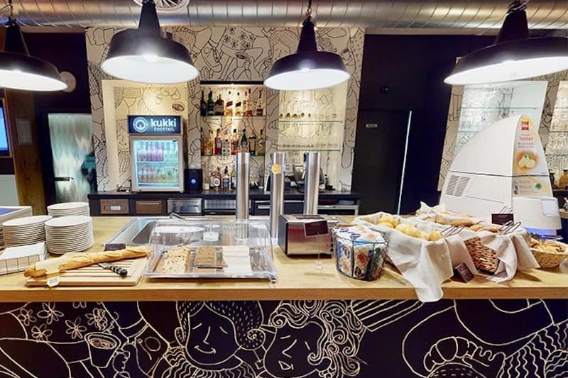 Buffettheke mit Baguette, Gebäck, Tellern und Kaffeemaschine vor einer Wand mit schwarzweißzeichnung und drei Hängelampen.