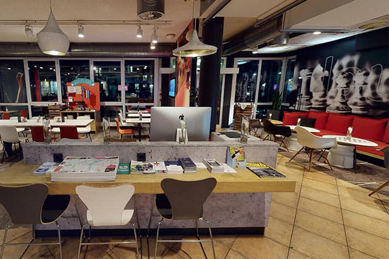 Hotellounge mit Tresen, Computer, ausliegenden Broschüren, Tischen und bunten Stühlen vor großen Fenstern.