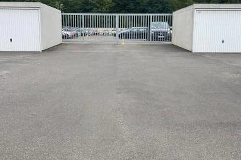 Große asphaltierte Fläche vor geschlossenem Metalltor zwischen zwei weißen Garagen, geparkte Autos hinter dem Tor sichtbar.