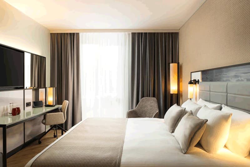 Helles Hotelzimmer mit Doppelbett, gepolstertem Kopfteil, Nachttischlampen, Schreibtisch mit Stuhl und großer Fensterfront mit Vorhängen.