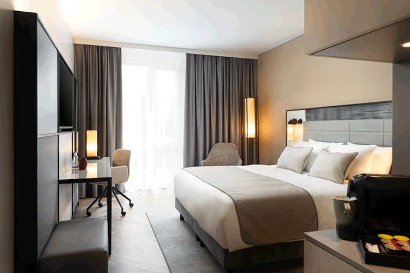 Helles Hotelzimmer mit Doppelbett, Schreibtisch, Sessel, bodentiefen Fenstern und Kaffeemaschine.