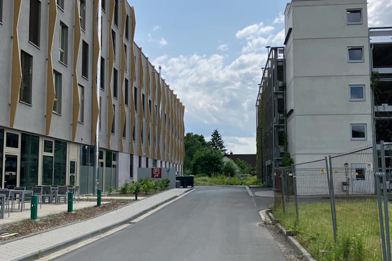 Straße zwischen modernem Hotelgebäude mit Außensitzbereichen links und eingezäuntem Parkhaus rechts unter leicht bewölktem Himmel.