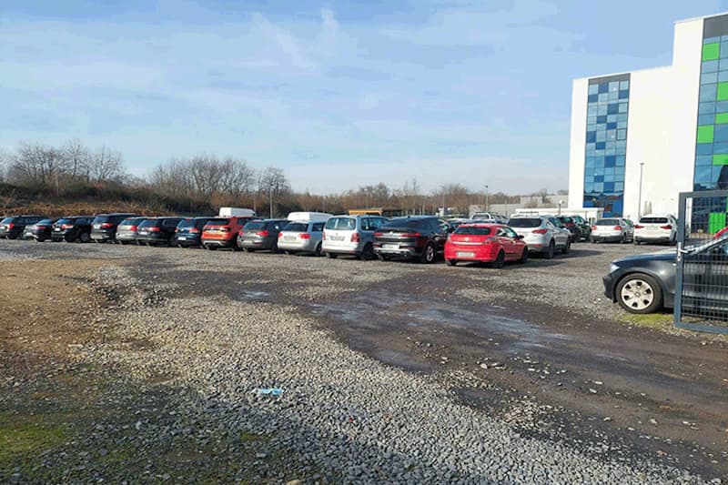 Schotterparkplatz mit mehreren geparkten Autos, Metallzaun und einem Gebäude mit blauen Fassadenplatten.