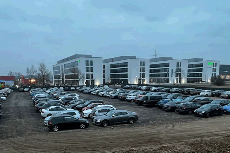 Großer geschotterter Parkplatz mit Reihen geparkter Autos vor modernen Bürogebäuden bei Dämmerung.