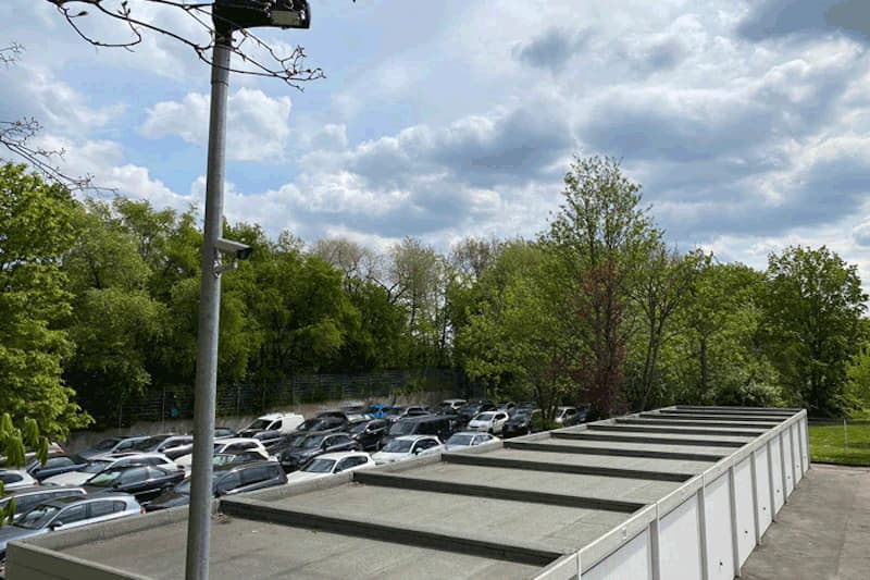 Parkplatz mit dicht geparkten Autos, Sicherheitskamera auf Mast und Garagenreihe vor Bäumen unter bewölktem Himmel.