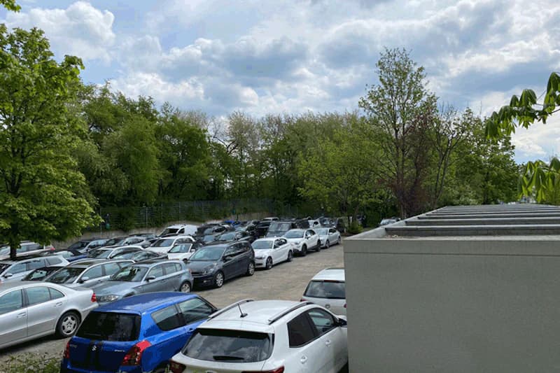 Außenparkplatz mit dicht geparkten Pkw, Bäumen und Zaun im Hintergrund unter bewölktem Himmel.