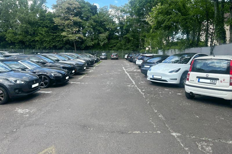 Parkplatz mit zwei Reihen geparkter Autos auf Asphalt, Maschendrahtzaun und Bäume im Hintergrund.
