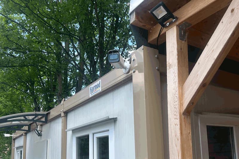 Überwachungskamera und Flutlicht an einem Modulcontainer mit Holzbalken und Bäumen im Hintergrund.