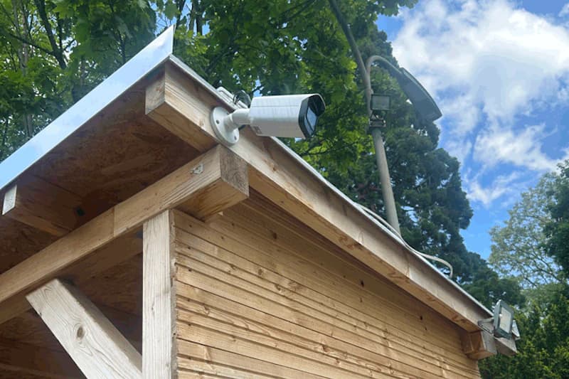 Weiße Überwachungskamera an der Dachkante eines Holzschuppens, daneben Scheinwerfer und Bäume im Hintergrund.