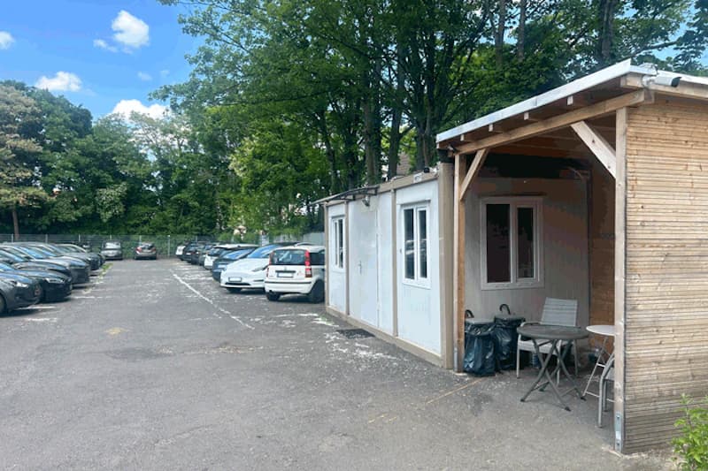 Asphaltierter Parkplatz mit Reihen geparkter Autos, Containerbüro und überdachte Sitzecke rechts, Bäume und Zaun im Hintergrund.