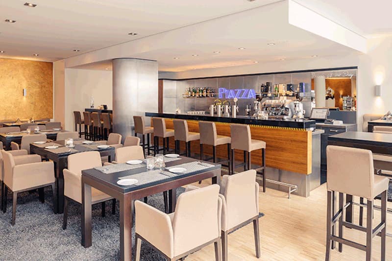 Innenraum eines Hotelrestaurants mit Tischen, beigen Stühlen und einer Theke mit Neonaufdruck PIAZZA.