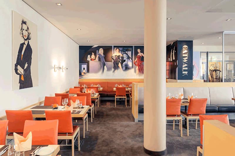 Hotelrestaurant mit gedeckten Tischen, orangefarbenen Stühlen, Säulenpfeiler und Modebildern an der Wand.