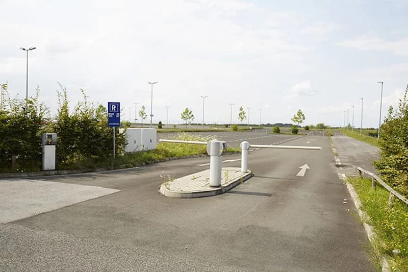 Zufahrt mit Schranke zu einem fast leeren Parkplatz mit blauem P-Schild und Laternen.