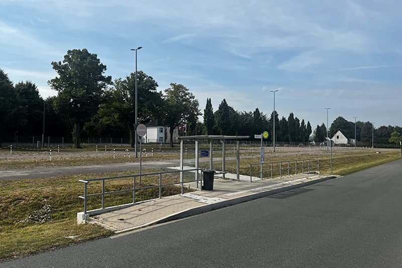 Überdachte Wartehalle mit Müllbehälter und Geländer an Straße vor offenem Parkplatz und Baumreihe.