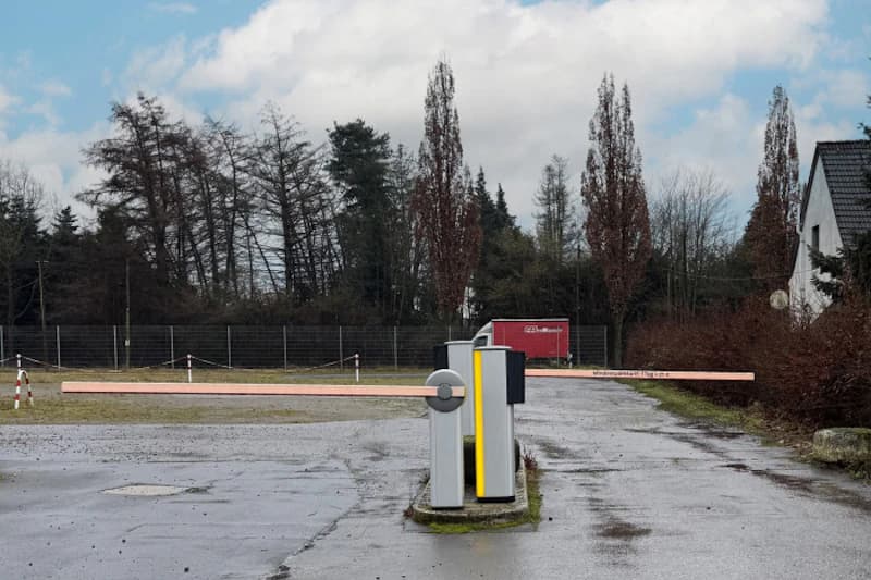 Schranke in Einfahrt zu leerem Parkplatz mit nassem Belag, Büschen, Bäumen und rotem Anhänger im Hintergrund.