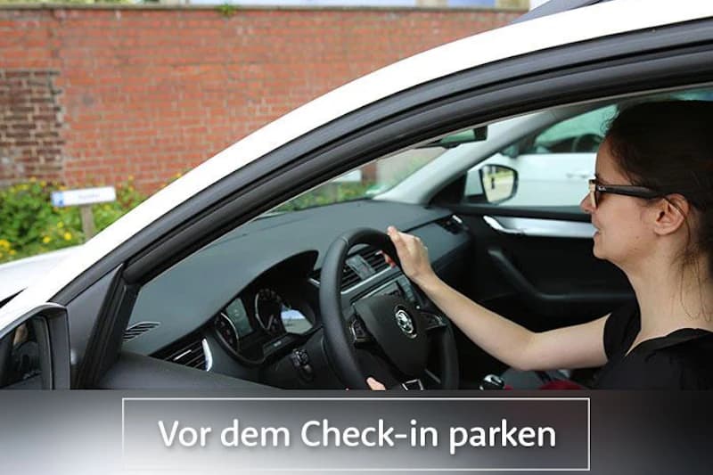 Frau im Fahrersitz eines weißen Autos hält das Lenkrad und schaut nach vorn; Backsteinmauer im Hintergrund.