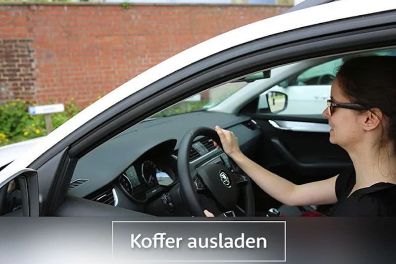 Frau sitzt am Steuer eines weißen Autos, Hand am Lenkrad, Blick nach vorn; Schriftzug Koffer ausladen sichtbar.
