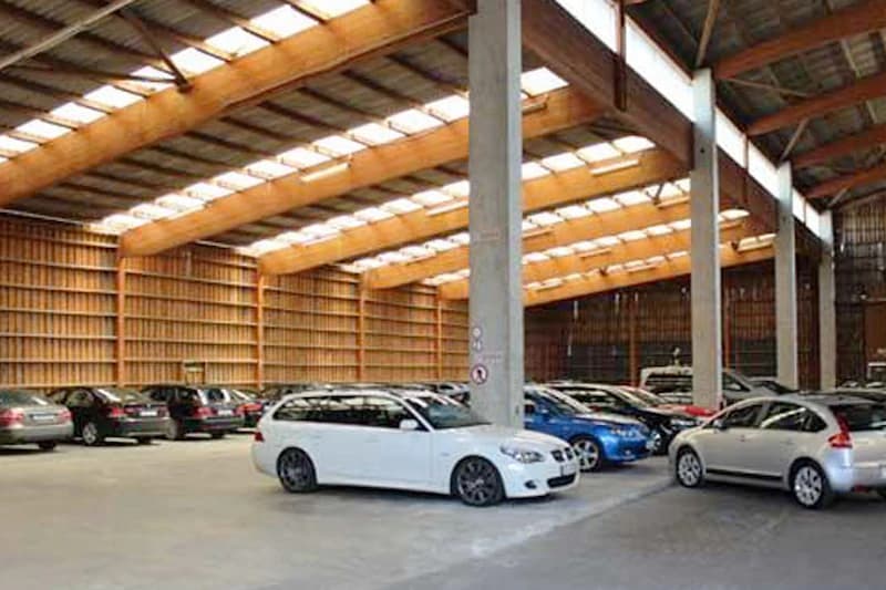 Große Parkhalle mit geparkten Autos, Holzbalken und Betonpfeilern unter Tageslichtfenstern.