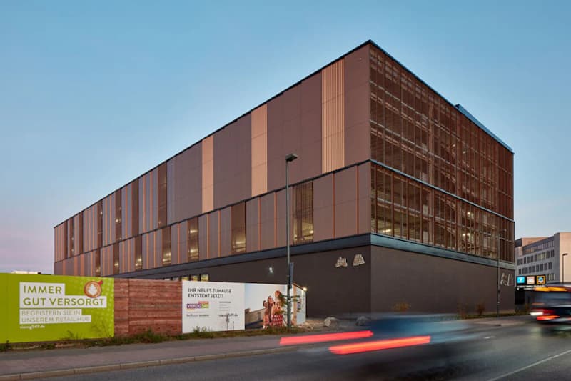 Mehrstöckiges Parkhaus mit Metallverkleidung und beleuchteten Rampen, Bauzaun und Werbebanner an der Straße.