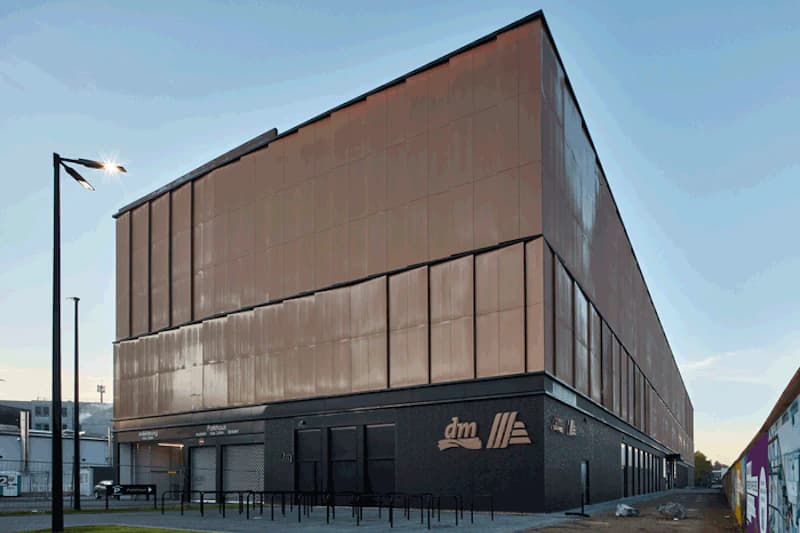 Mehrstöckiges Parkhaus mit bronzefarbener Metallfassade, Schriftzug dm am Sockel, Lichtmasten und Fahrradständern.