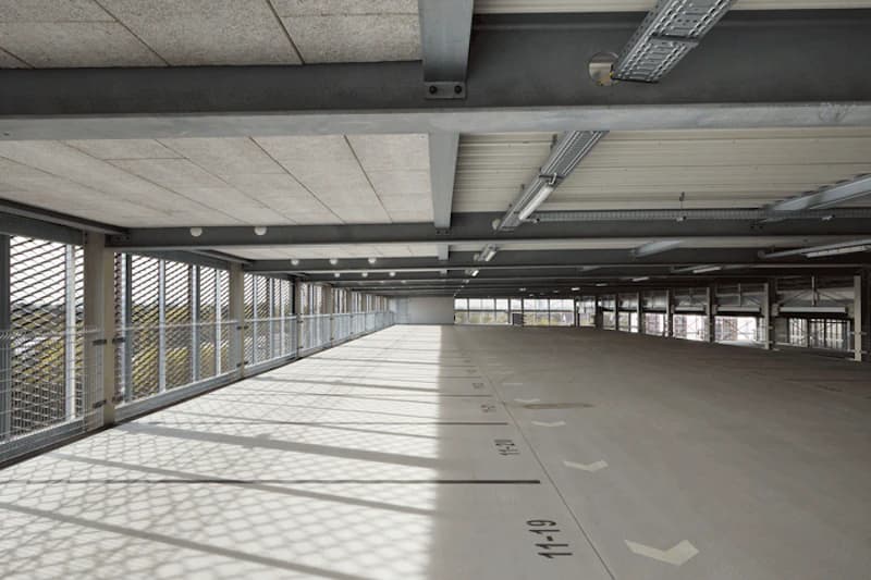 Leeres oberes Parkdeck mit markierten Stellplaetzen, Metallgitter an den Seiten, Stahltraegern und Deckenlampen.