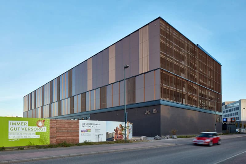 Mehrstöckiges Parkhaus mit Metallfassade, Bauzaun mit Werbetafeln und Straße mit fahrendem roten Auto.