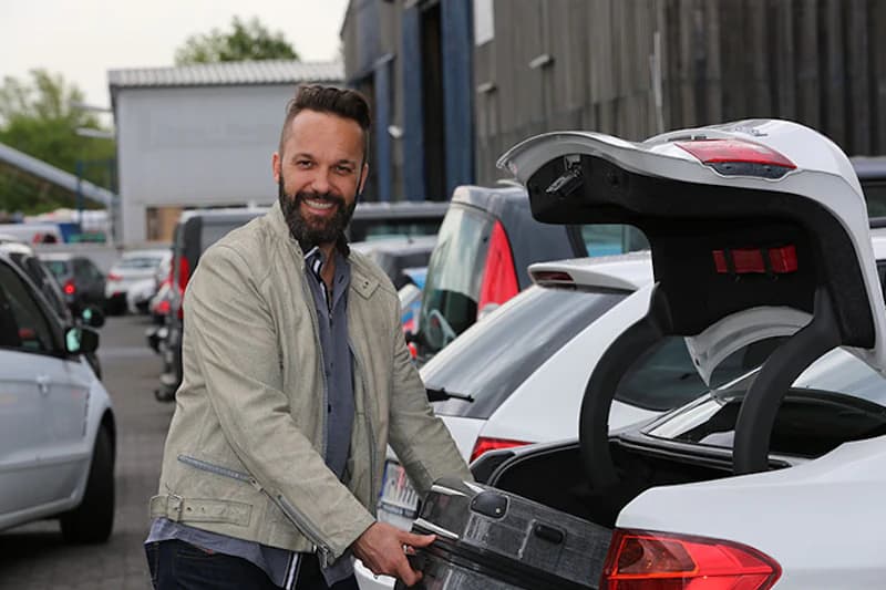 Mann lädt Koffer in den offenen Kofferraum eines weißen Autos auf einem Parkplatz mit mehreren Fahrzeugen.