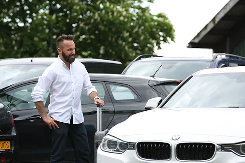 Mann mit Koffer neben weißem BMW auf Freiflächenparkplatz mit weiteren geparkten Fahrzeugen.