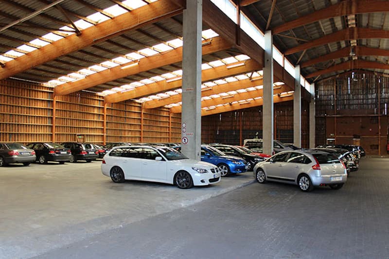 Große Innenparkhalle mit Reihen geparkter Autos, Holzträgern, Betonstützen und Lichtbändern im Dach.