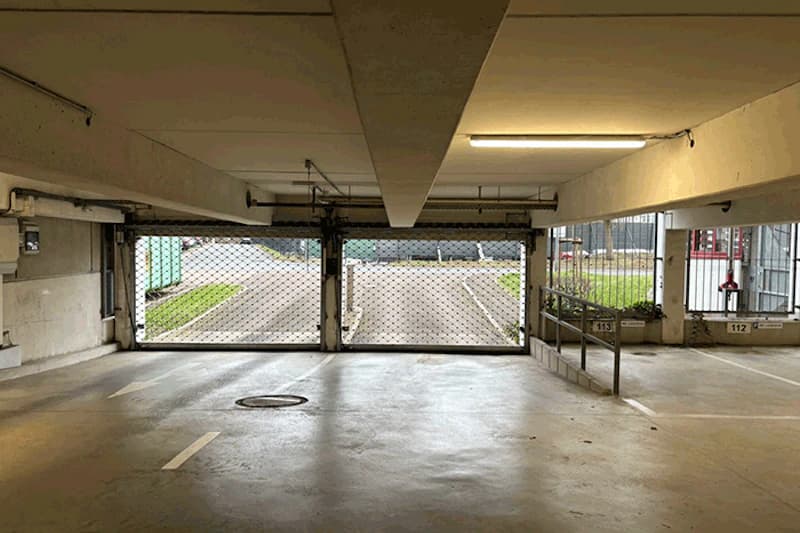 Beleuchtetes Parkdeck mit heruntergelassenen Rollgittern an der Ausfahrt, markierten Stellplätzen und Betondecke.