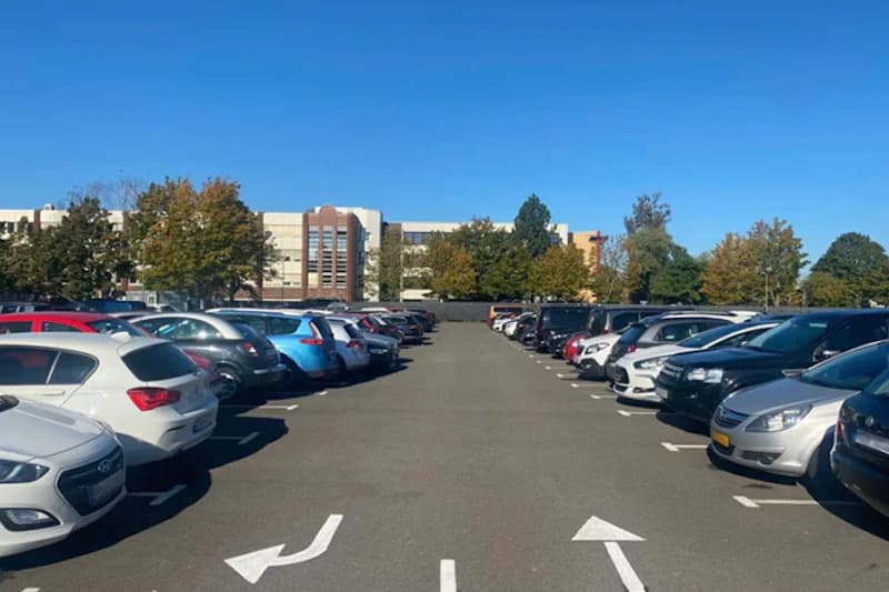 Reihen geparkter Autos auf Außenparkplatz, Fahrbahnmarkierungen mit Pfeilen, Gebäude und Bäume im Hintergrund.